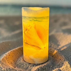 May include: A cylindrical pillar candle with a warm yellow glow, featuring a printed graphic of a large seashell resting on a sandy beach with ocean waves in the background. The surface of the candle is smooth and illuminated from within.