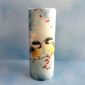 May include: A cylindrical candle with a winter scene featuring two small birds perched on a branch with red berries. The candle has a light blue and white background with a watercolor effect.