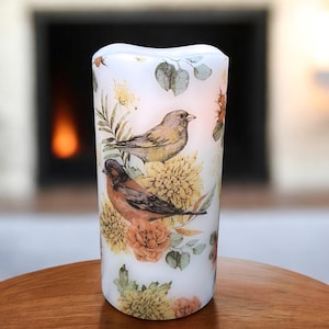 May include: A white pillar candle featuring a floral and bird design. The candle has a curved top and is decorated with illustrations of two birds, yellow flowers, and green leaves. The candle is designed to look like it is lit.