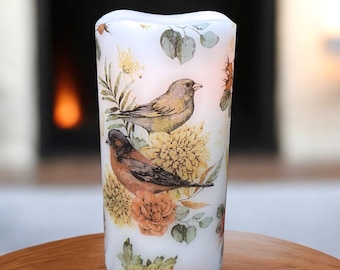 May include: A white pillar candle featuring a floral and bird design. The candle has a curved top and is decorated with illustrations of two birds, yellow flowers, and green leaves. The candle is designed to look like it is lit.