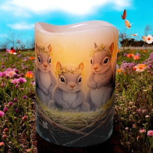 May include: A candle with a design of three squirrels wearing flower crowns sitting in a nest. The candle is white and has a warm glow.