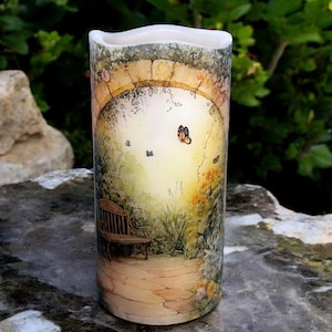 May include: A cylindrical candle with a watercolor design of a garden scene. The scene features a stone archway, a bench, and butterflies flying through the air. The candle is lit and the light shines through the archway.