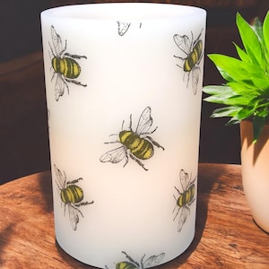 May include: A cylindrical, off-white candle holder with a repeating pattern of illustrated bees in black and yellow. The candle holder is on a wooden surface, with a potted plant visible in the background.