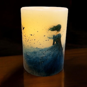 May include: A white pillar candle with a blue and white image of a woman with long hair blowing in the wind. The woman is wearing a blue dress and is walking away from the viewer. There are small birds flying around her.