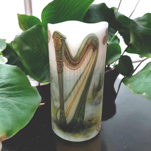 May include: A white pillar candle with a green and gold Celtic harp design. The harp is surrounded by a green and gold Celtic knot design.