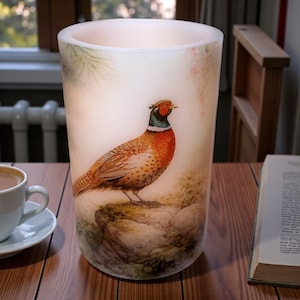May include: A cylindrical, frosted glass candle holder featuring a detailed illustration of a pheasant. The bird is depicted with vibrant orange, brown, and green plumage, perched on a rock. The background has soft, muted tones.