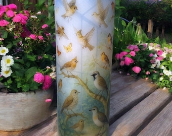May include: A cylindrical decorative lamp featuring a painted design of various birds perched on branches and flying against a light blue background. The lamp is placed outdoors near potted flowers and greenery.