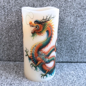 May include: A white pillar candle with a colorful image of a dragon wrapped around it. The dragon is orange, green, and yellow with a detailed design.