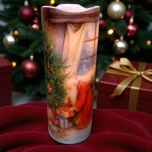 May include: A cylindrical LED candle featuring a printed illustration of Santa Claus in a red robe kneeling beside a decorated Christmas tree and wrapped gifts. The scene is set indoors near a window with a view of a snowy landscape outside.