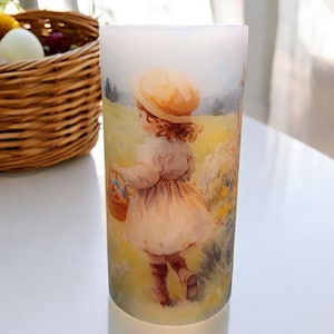 May include: A white pillar candle with a colorful image of a girl in a straw hat holding a basket of Easter eggs. The girl is walking through a field of flowers.