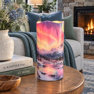 May include: A cylindrical LED candle featuring a vibrant print of the aurora borealis over a snow-covered mountain landscape with a reflective lake. The candle has a wavy top edge and emits a warm, soft glow from within the illuminated surface.