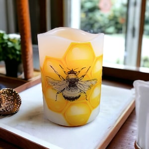 May include: A white pillar candle with a yellow honeycomb design and a black and white illustration of a bee. The candle is lit and glowing.