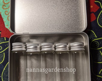 Puede incluir: Una caja rectangular de metal plateado que contiene cinco viales de vidrio transparente vacíos con tapas de rosca plateadas. El texto nannasgardenshop está impreso en blanco en el centro de los viales de vidrio dentro del recipiente abierto.