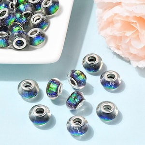 May include: A collection of iridescent glass beads with a hole through the center. The beads have a clear base with colorful, swirling patterns of blue, green, and purple. Some beads are scattered on a light blue surface, while others are in a white dish.