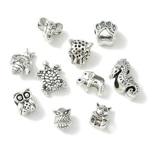 May include: A collection of ten silver-toned metal beads shaped like various animals and nature elements, including an elephant head, a paw print, a leopard head, a bee, a turtle, an elephant, a seahorse, two owls, and a small bear figure.
