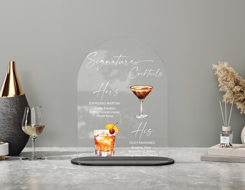 May include: Clear acrylic sign with the words "Signature Cocktails" and drink names "Hers" and "His". The "Hers" cocktail is an Espresso Martini, and the "His" cocktail is an Old Fashioned. The sign is on a black base.