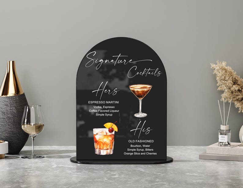 May include: A black arched sign with the words "Signature Cocktails" in white script. The sign lists two cocktail recipes: Espresso Martini and Old Fashioned, with illustrations of each drink. The sign is on a black base.