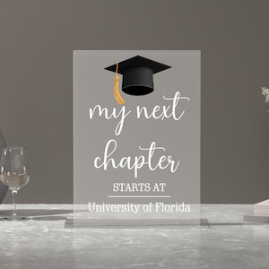 May include: A clear acrylic sign with the text "my next chapter starts at University of Florida" and a black graduation cap with a gold tassel.