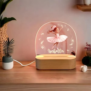 以下が含まれることがあります： バレリーナのイラストと「イザベラ」という名前が描かれた透明なアクリルパネルが付いた木製ナイトライト。温白色の光で照らされています。