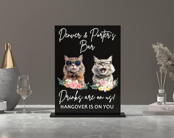 Op de afbeelding: Zwart acryl bord met de tekst "Denver & Porter's Bar" en twee katten met zonnebrillen en bloemmotieven. De tekst onder de katten luidt "Drinks are on us! Hangover is on you."