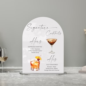 May include: A white arched sign with the words "Signature Cocktails" in elegant script. The sign features two cocktail recipes: "Hers" (Espresso Martini) and "His" (Old Fashioned), with illustrations of each drink. The sign is on a silver stand.