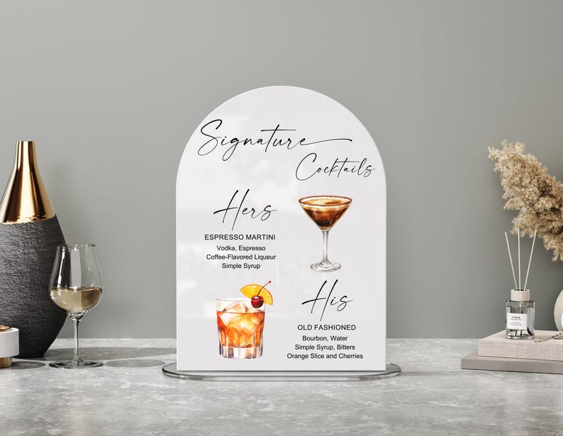 May include: A white arched sign with the words "Signature Cocktails" in elegant script. The sign features two cocktail recipes: "Hers" (Espresso Martini) and "His" (Old Fashioned), with illustrations of each drink. The sign is on a silver stand.