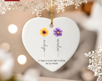 Op de afbeelding: Witte hartvormige keramische hanger met twee bloemen, een gele en een paarse, met de namen "evelyn" en "amelia" eronder. De hanger heeft de tekst "Het duurt lang om een ​​oude vriend te laten groeien."