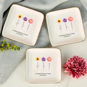Op de afbeelding: Drie witte keramische sieradenbakjes met gouden rand. Elk bakje heeft een bloemmotief met drie bloemen en de namen "Emma", "Amelia" en "Evelyn" eronder. De tekst "Als vrienden bloemen waren, zou ik je kiezen" staat op elk bakje gedrukt.