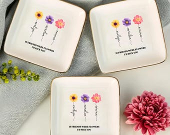 Op de afbeelding: Drie witte keramische sieradenbakjes met gouden rand. Elk bakje heeft een bloemmotief met drie bloemen en de namen "Emma", "Amelia" en "Evelyn" eronder. De tekst "Als vrienden bloemen waren, zou ik je kiezen" staat op elk bakje gedrukt.