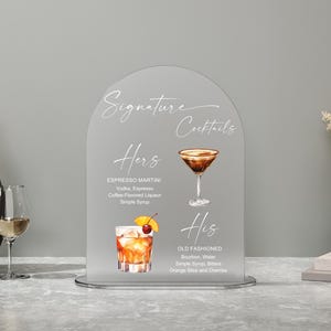May include: A frosted acrylic sign with the words "Signature Cocktails" in elegant script. The sign features illustrations and ingredients for "Hers" Espresso Martini and "His" Old Fashioned cocktails, perfect for a wedding or event.