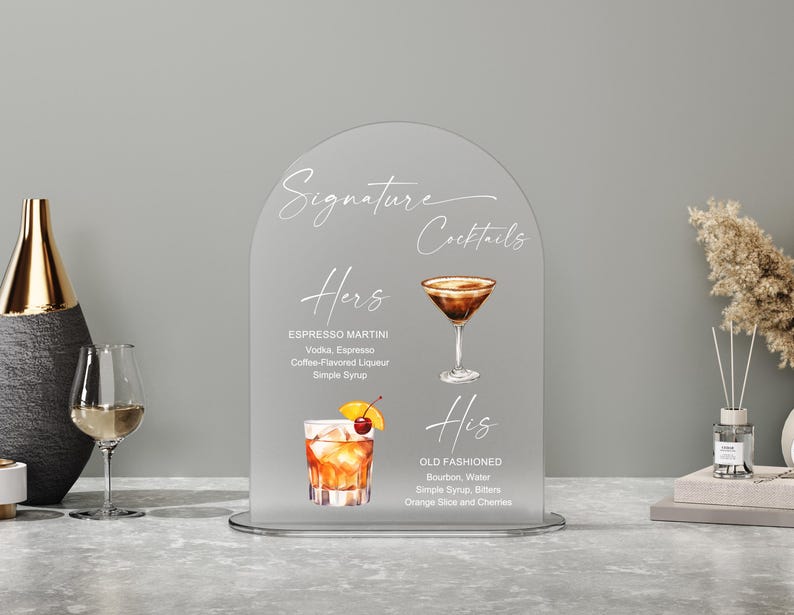 May include: A frosted acrylic sign with the words "Signature Cocktails" in elegant script. The sign features illustrations and ingredients for "Hers" Espresso Martini and "His" Old Fashioned cocktails, perfect for a wedding or event.