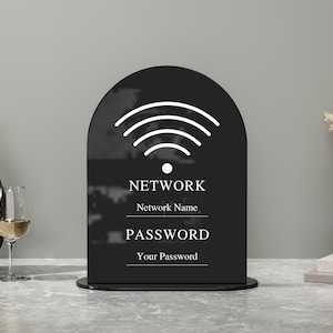 Op de afbeelding: Een zwart acrylen WiFi-wachtwoordschild met een afgeronde bovenkant. Het bord toont een wit WiFi-symbool en de woorden "NETWERK", "WACHTWOORD" en velden voor netwerknaam en wachtwoord. Het bord staat op een grijs oppervlak.