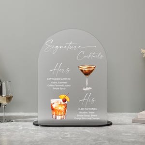 May include: A clear acrylic sign with an arched top displays "Signature Cocktails" in elegant script. The sign features illustrations and ingredients for "Hers" Espresso Martini and "His" Old Fashioned cocktails, with a black base.