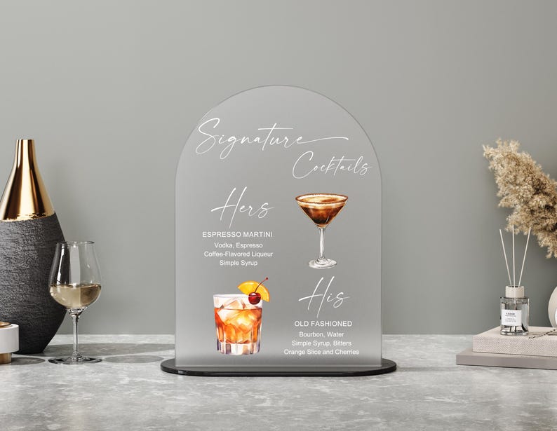 May include: A clear acrylic sign with an arched top displays "Signature Cocktails" in elegant script. The sign features illustrations and ingredients for "Hers" Espresso Martini and "His" Old Fashioned cocktails, with a black base.