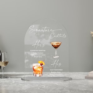 May include: Clear acrylic sign with the words "Signature Cocktails" and drink recipes for "Hers" and "His." The "Hers" cocktail is an Espresso Martini, and the "His" cocktail is an Old Fashioned. The sign is on a table with a vase and a wine glass.