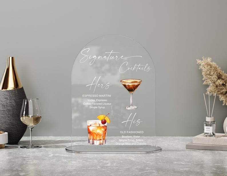 May include: Clear acrylic sign with the words "Signature Cocktails" and drink recipes for "Hers" and "His." The "Hers" cocktail is an Espresso Martini, and the "His" cocktail is an Old Fashioned. The sign is on a table with a vase and a wine glass.