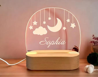 May include: A wooden night light with a clear acrylic panel featuring a crescent moon, clouds, and stars. The name "Sophia" is engraved on the panel. The light is illuminated with a warm white glow.