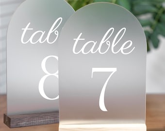 May include: Two frosted acrylic table number signs with wooden bases. The sign on the left says "table 8" and the sign on the right says "table 7".