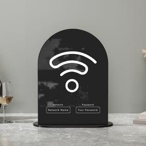 May include: A black acrylic WiFi sign featuring a white WiFi symbol. The sign has a rounded top and a rectangular base. It includes fields for network name and password. Decorative items such as a vase, wine glass, and dried flowers are in the background.