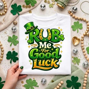 Puede incluir: Camiseta blanca con un diseño del Día de San Patricio. El diseño presenta un sombrero verde de duende, una herradura y el texto "Rub Me For Good Luck" en verde y amarillo. La camiseta está rodeada de tréboles y cuentas decorativas.