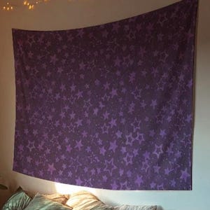 Puede incluir: Un gran tapiz morado con un patrón de estrellas repetidas cuelga de una pared. Las estrellas son de un tono morado más claro. El tapiz está sobre una cama con almohadas y una colcha. Luces de cadena están arriba.