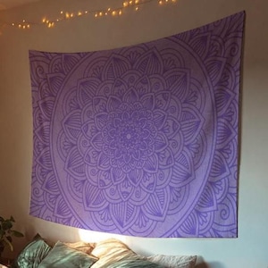 May include: A large, lavender mandala tapestry hanging on a wall above a bed. The tapestry features a detailed, symmetrical floral design. The room has a cozy atmosphere with pillows and a string of lights.