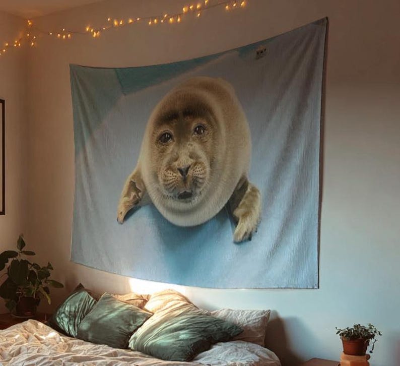 May include: A large wall tapestry featuring a realistic image of a seal. The seal is light brown with a detailed face and is set against a light blue background. The tapestry is hanging above a bed with pillows.