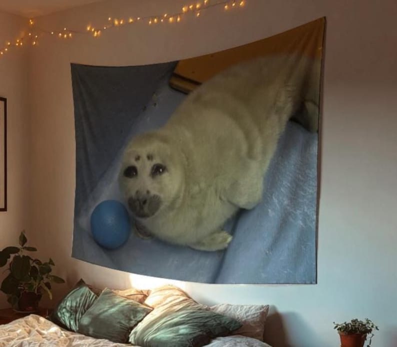 May include: A large wall tapestry featuring a light-colored seal with dark eyes and a blue ball. The seal is lying on a blue surface. The tapestry is hanging on a wall with string lights.