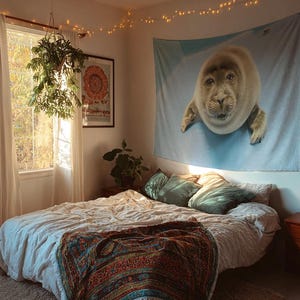 May include: A bedroom scene featuring a bed with a white comforter and a colorful patterned blanket. A large tapestry of a seal hangs on the wall above the bed. The room is decorated with plants and string lights.