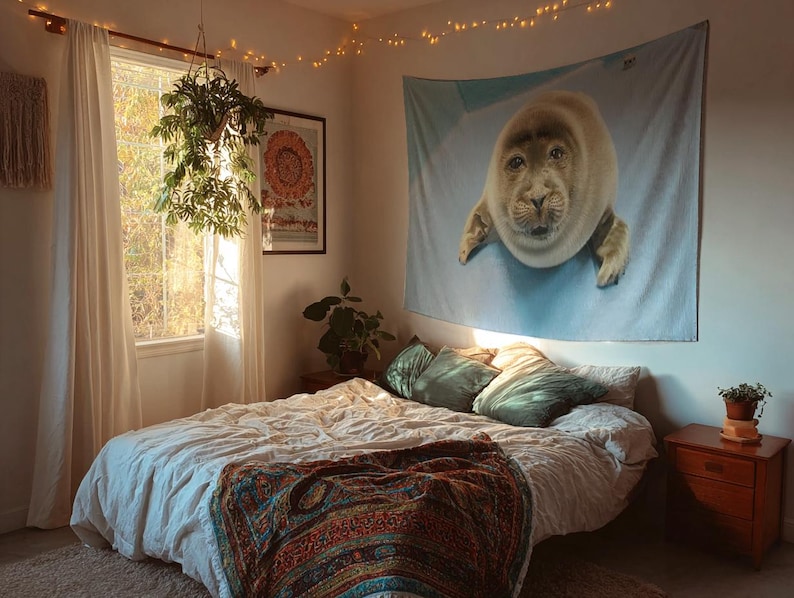 May include: A bedroom scene featuring a bed with a white comforter and a colorful patterned blanket. A large tapestry of a seal hangs on the wall above the bed. The room is decorated with plants and string lights.
