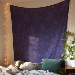 Puede incluir: Un gran tapiz morado oscuro con un patrón floral cuelga sobre una cama. El tapiz es el punto focal, con un efecto degradado y un borde superior ligeramente curvado. También se ven una almohada verde y una planta en una maceta blanca.