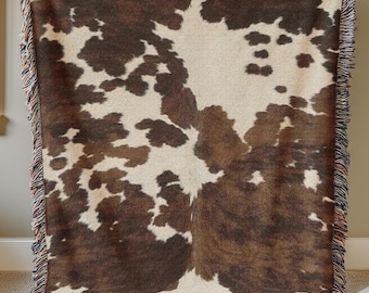 Op de afbeelding: Een geweven sprei met een koeienhuidpatroon in bruin- en crèmetinten. De deken heeft een gefranjerde rand met een mix van kleuren. De deken wordt door een persoon vastgehouden.