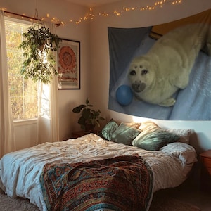 May include: A cozy bedroom scene with a large bed covered in a white comforter and a colorful patterned blanket. A tapestry of a seal hangs on the wall above the bed. The room is lit by natural light and string lights.