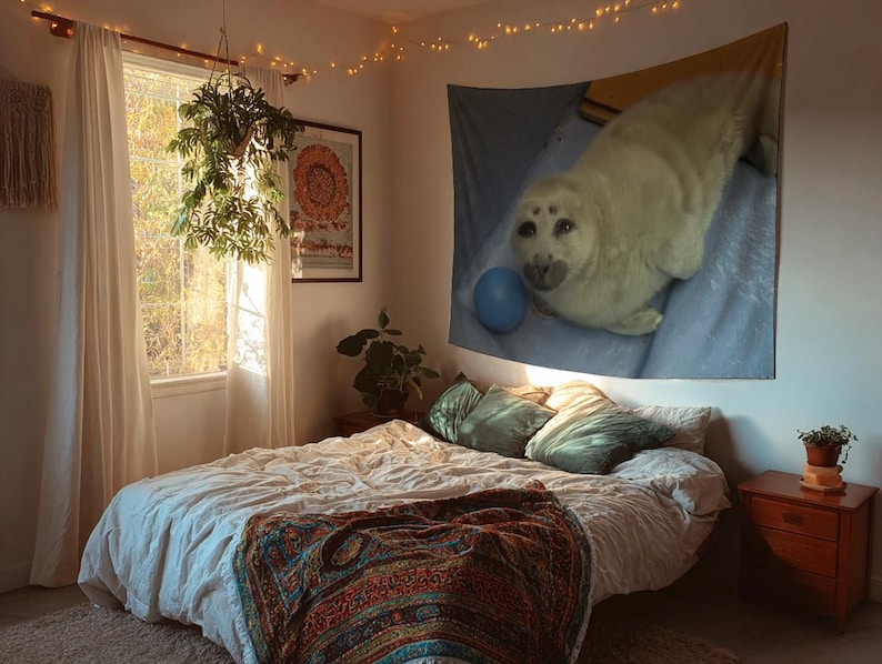 May include: A cozy bedroom scene with a large bed covered in a white comforter and a colorful patterned blanket. A tapestry of a seal hangs on the wall above the bed. The room is lit by natural light and string lights.